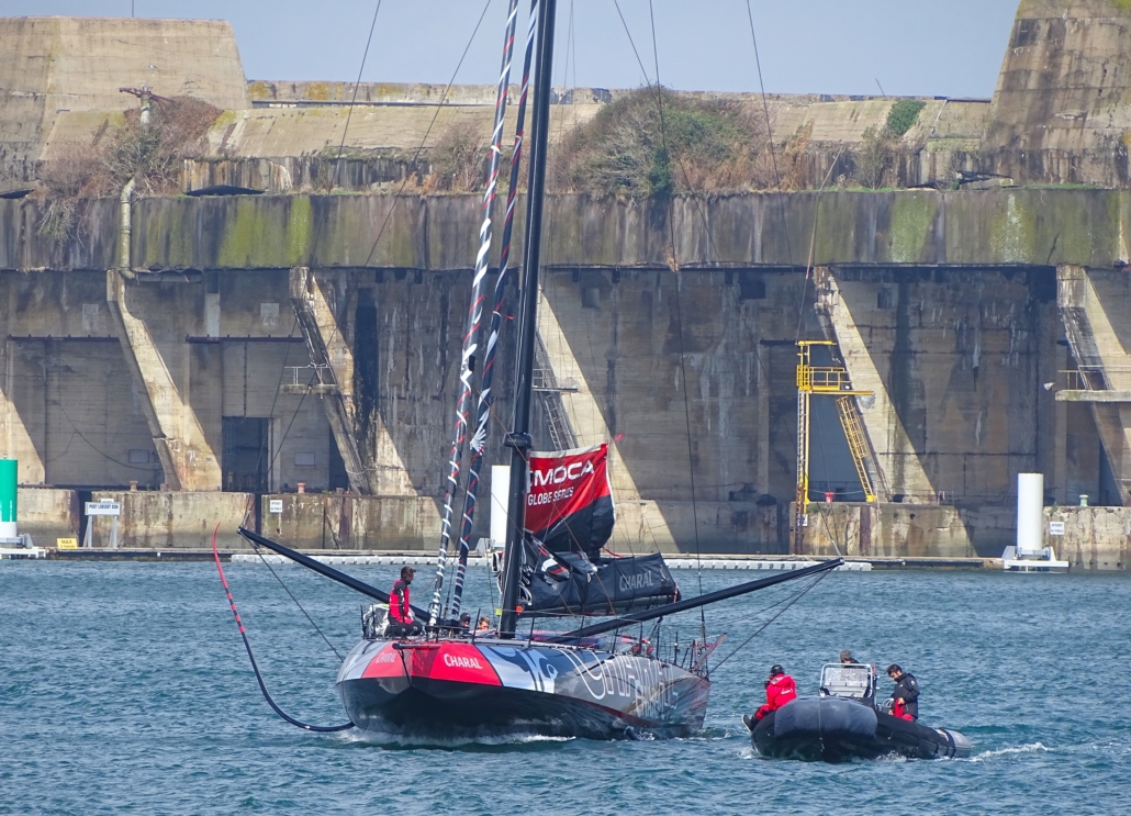 A racing yacht sailing out to sea, with a backdrop of  WW2 u-boat pens in Lorient