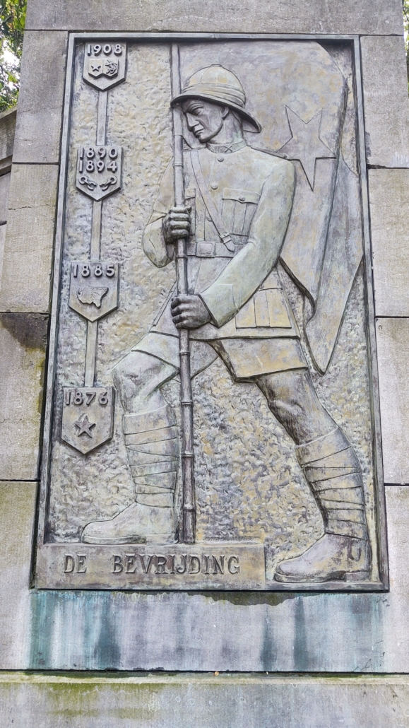 From the Leopold statue in Hasselt. On the right panel with the legend “DE BEVRIJDING” [The Liberation], we see a colonial officer with the Congolese flag in his hand.
To its left are four coats of arms, with dates referring to the following historical facts:
 •1876 and the Congolese flag: refers to the founding of the International African Association, which set itself the objective of exploring the basin of Congo
• 1885 and the map of the Congo Basin: proclamation of the International Congolese Association as a sovereign state, later Congo Independent State;
• 1890 – 1894 and a broken chain, symbolizing the campaign against the slave trade;
• 1908, the Congolese flag and the Belgian coat of arms: the annexation of the Congo to Belgium.