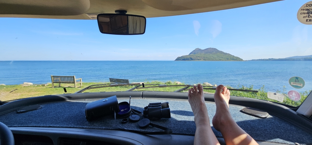 Feet on motorhome dashboard with view of sea and an island through the windscreen