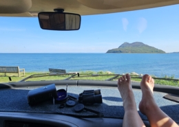 Feet on motorhome dashboard with view of sea and an island through the windscreen