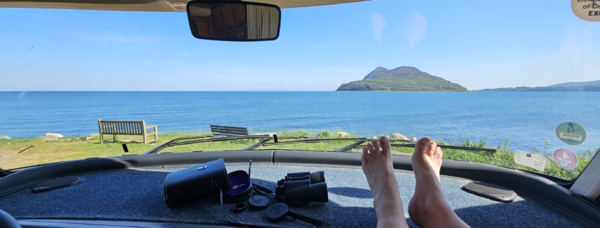 Feet on motorhome dashboard with view of sea and an island through the windscreen